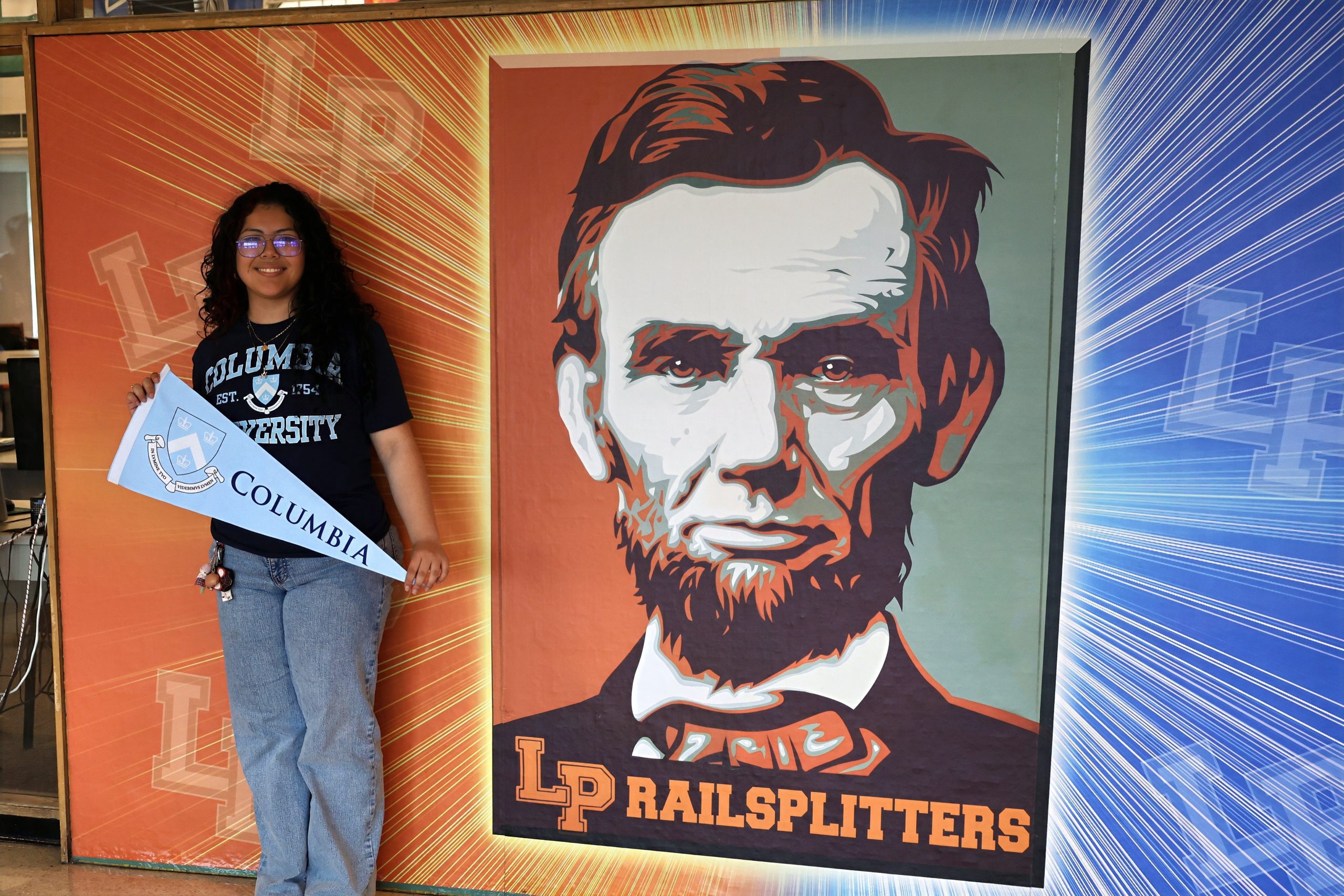 Smiling student with curly hair and glasses holds a blue'Columbia University' sash beside a large portrait of a man.