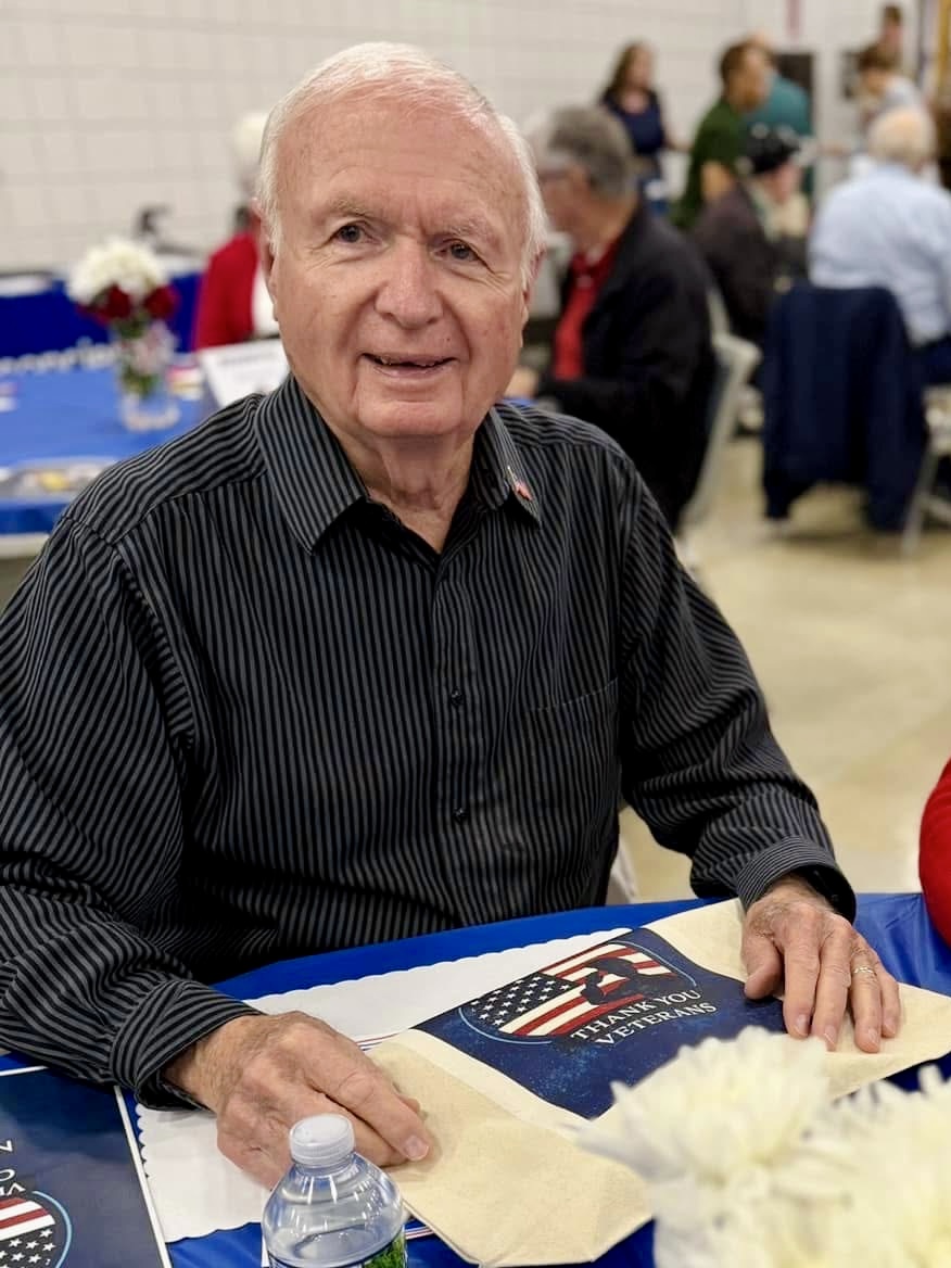 Older man in a black striped shirt sits at a table with a patriotic 'Thank you Veterans' napkin and a water bottle in front of him at a community event.