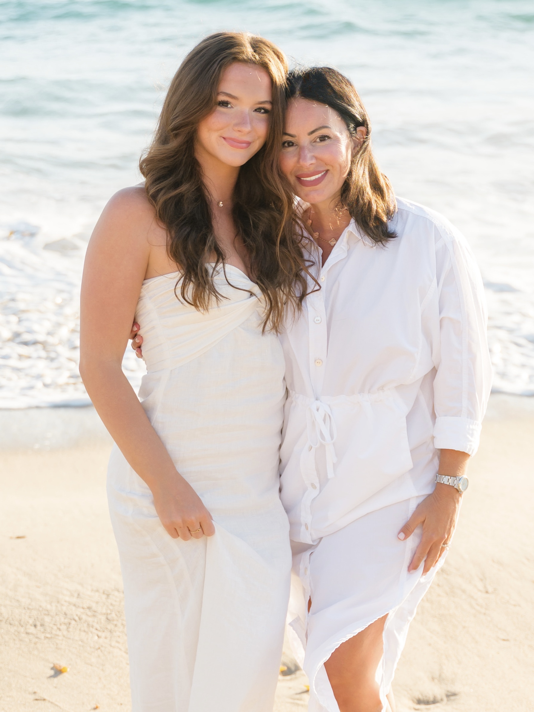 Two women in white outfits posing on a sunny beach, smiling at the camera.