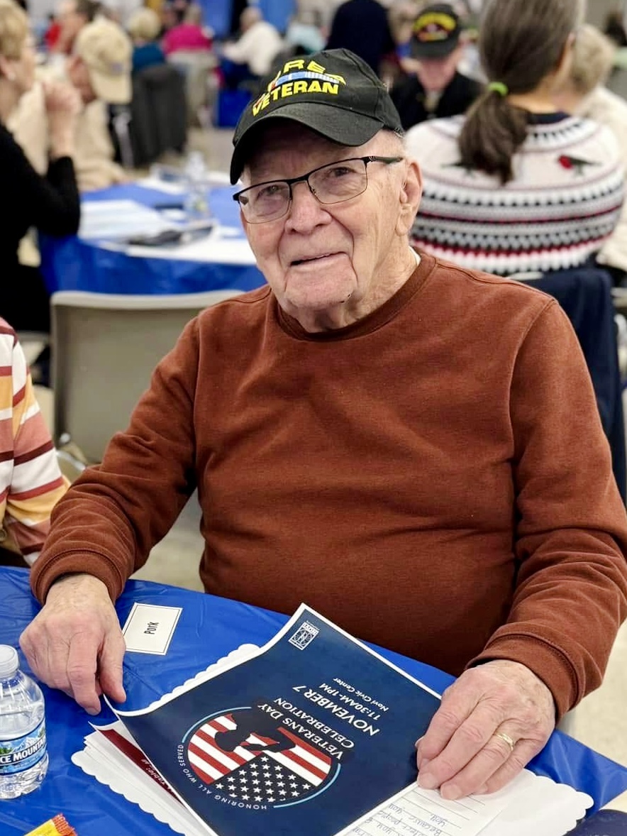 Older man in a brown sweater and a black cap that says 'VETERAN' sits at a blue-clothed table at a veterans event, with a patriotic brochure in front of him.