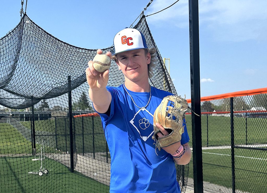Young baseball player in a blue jersey holds up a baseball toward the camera, wearing a white cap with red GC lettering, behind a protective netting.