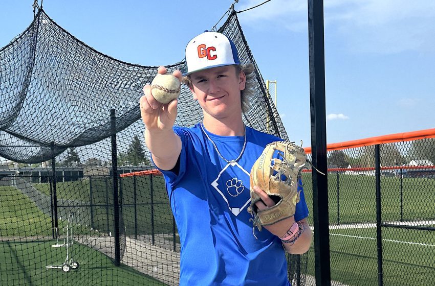 Young baseball player in a blue jersey holds up a baseball toward the camera, wearing a white cap with red GC lettering, behind a protective netting.