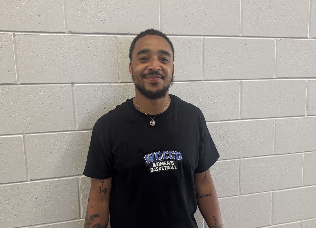 Young man with beard standing against a light gray cinder block wall, wearing a black shirt with 'WCCCD Women's Basketball' logo. Tattoos on both arms visible.