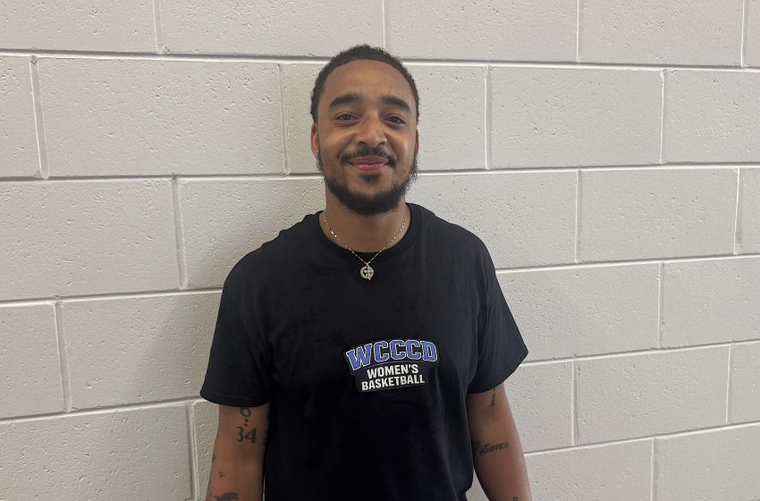Young man with beard standing against a light gray cinder block wall, wearing a black shirt with 'WCCCD Women's Basketball' logo. Tattoos on both arms visible.