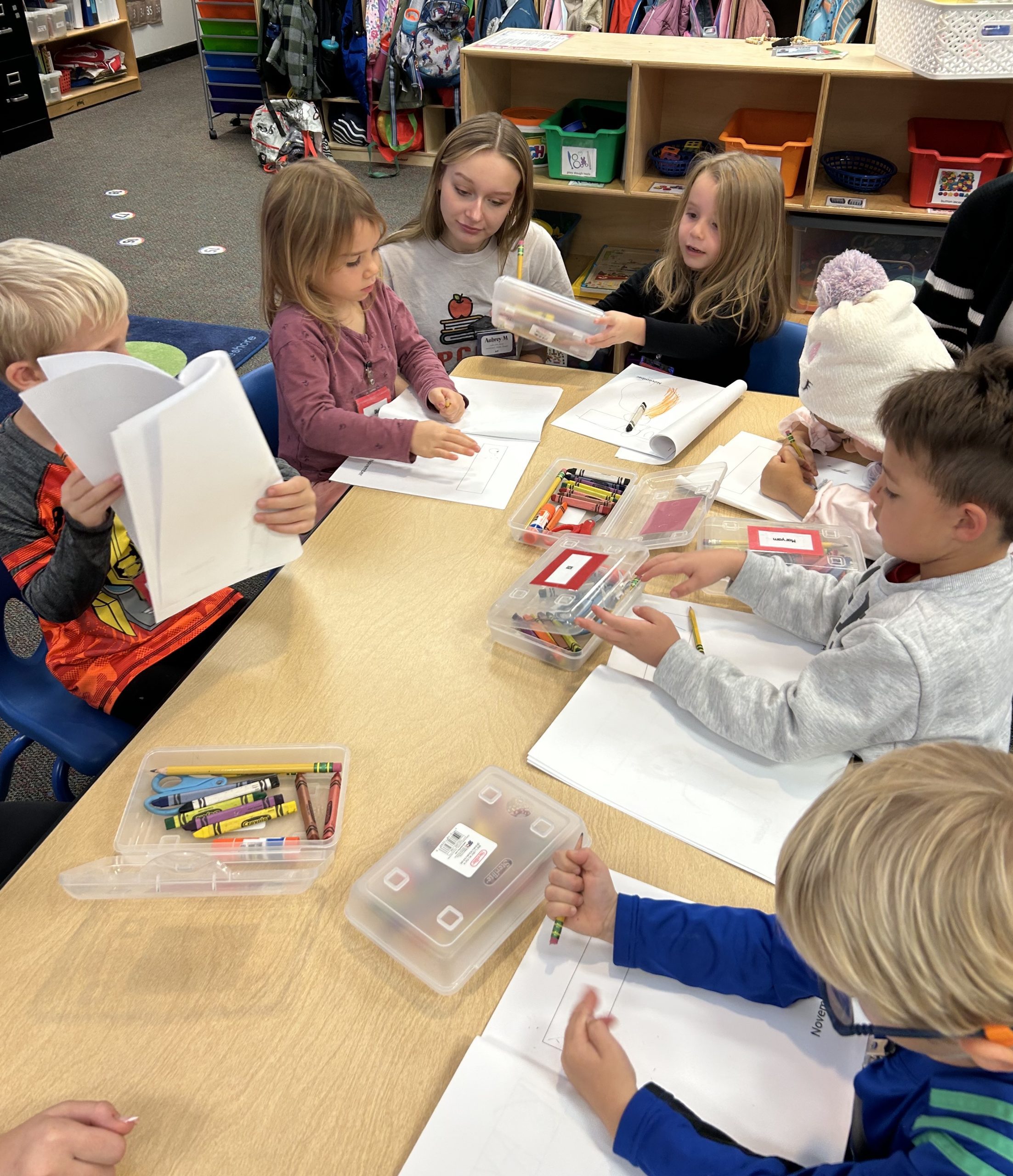 FutureTeachers2 - Social House News Children sit around a table in a classroom, drawing and coloring with crayons on sheets of paper.