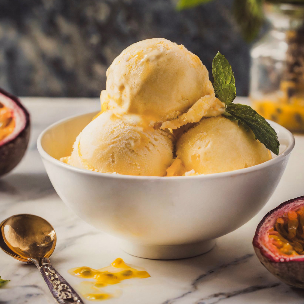 Three scoops of mango ice cream in a white bowl, garnished with mint leaves on a marble surface with passion fruit halves nearby.