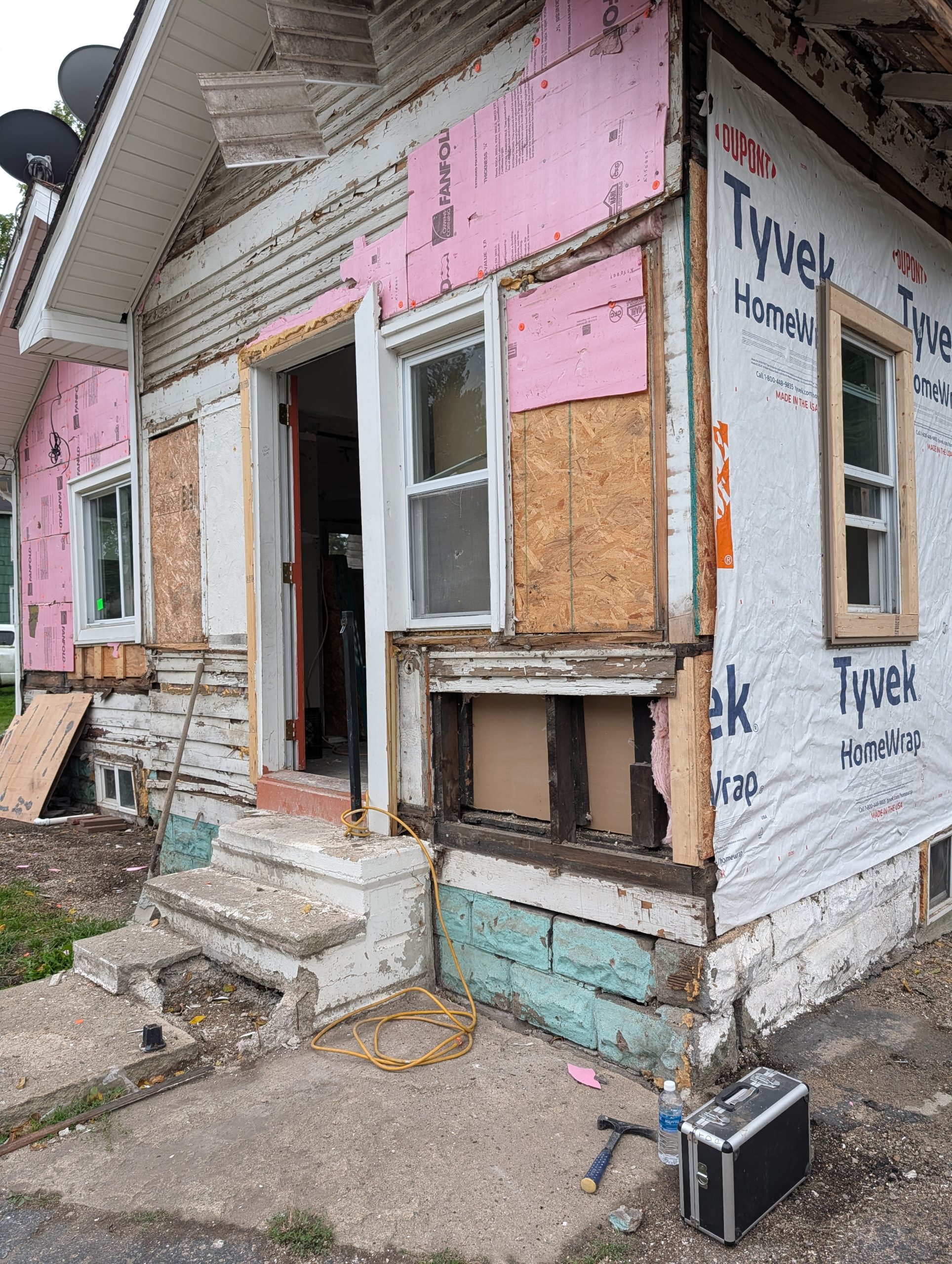 Grace Centers of Hope 85 Moreland 2 - Social House News Front corner of a house under renovation with pink insulation boards, Tyvek wrap, and peeling siding; tools and debris on the ground near an open doorway.