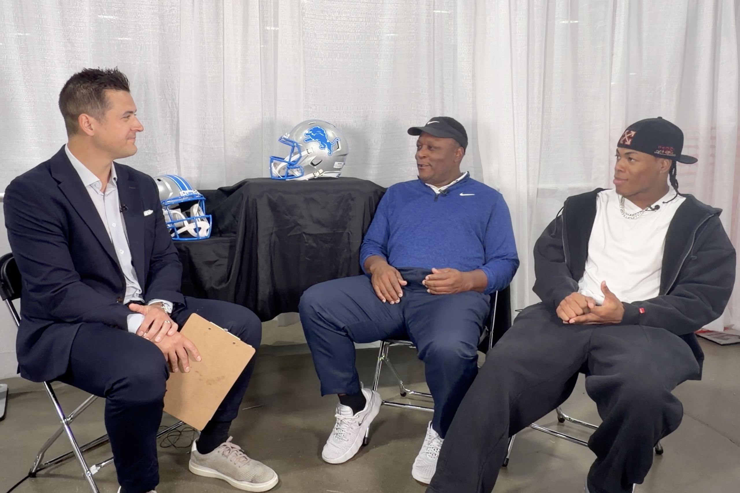 Three men sit in a row having a discussion; football helmets sit on a table behind them against a light curtain backdrop.