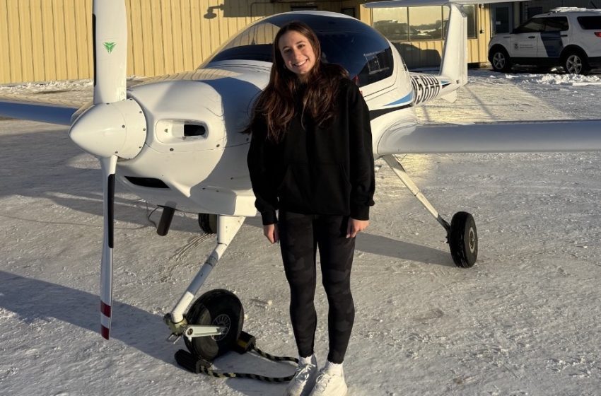 Smiling woman in a black hoodie stands beside a small white single-engine airplane on a snowy tarmac at Willow Run Airport.