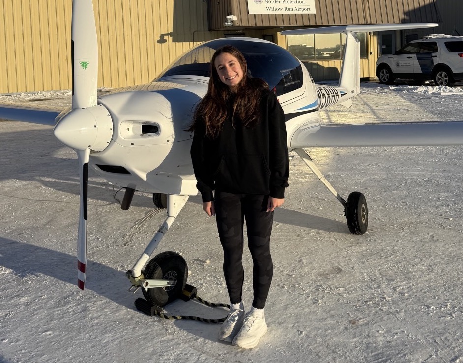 Smiling woman in a black hoodie stands beside a small white single-engine airplane on a snowy tarmac at Willow Run Airport.