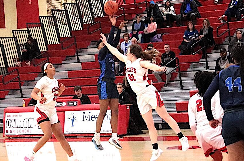 Girls' basketball game in progress on an indoor court; two white-jersey players leap for the ball while a blue-jersey defender jumps nearby, with spectators in red bleachers in the background.