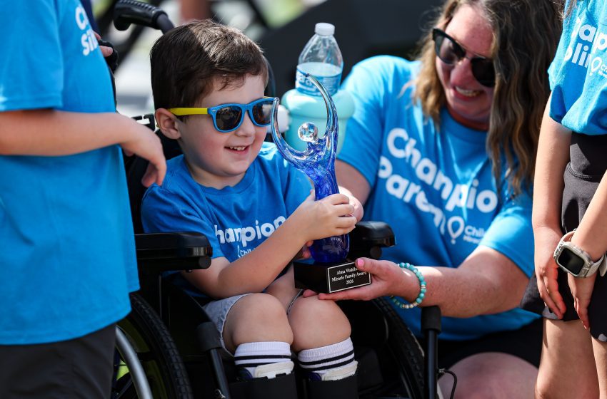 Boy in a wheelchair holding a blue glass trophy, smiling, with volunteers in blue shirts around him.