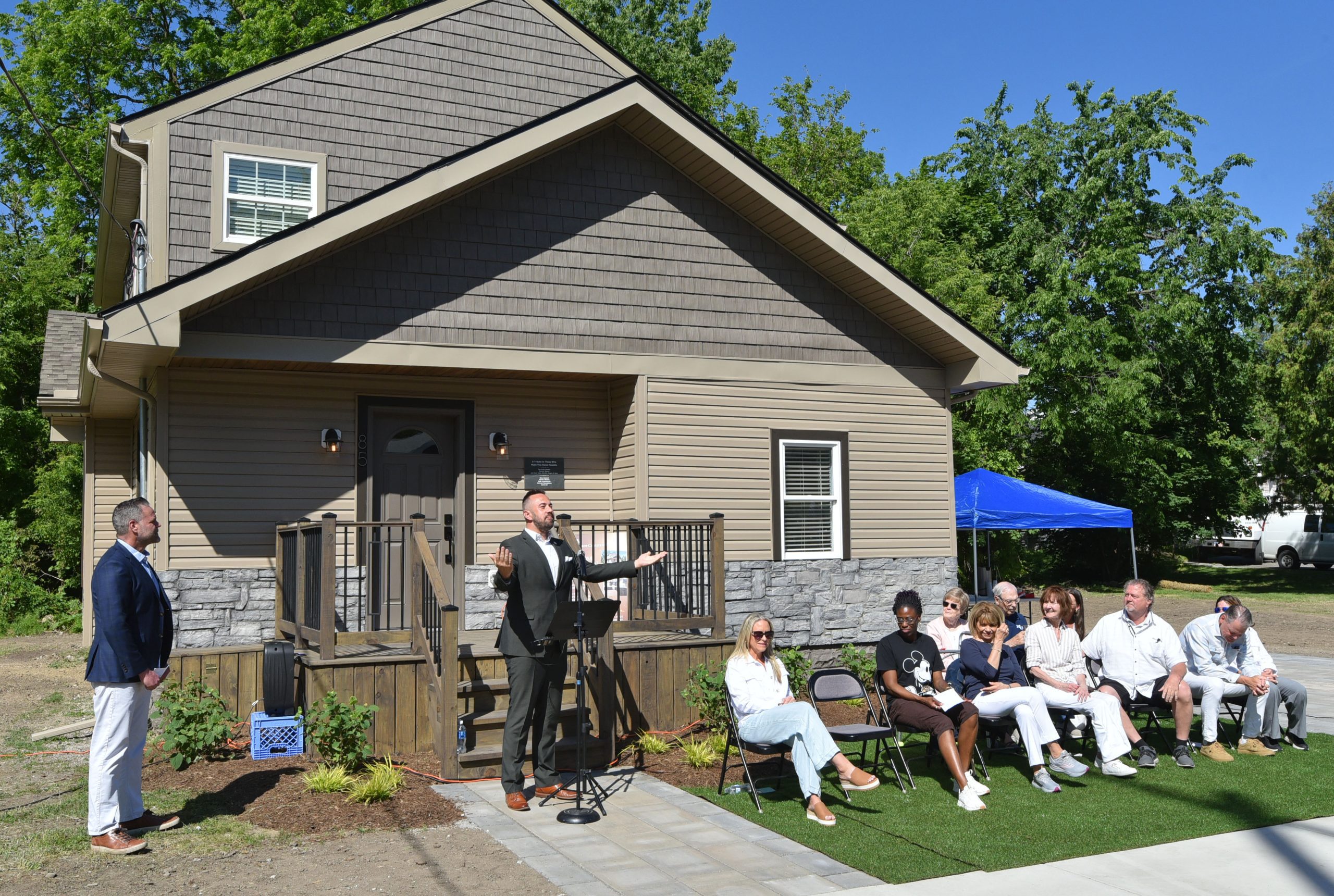 Man in a suit speaks into a microphone at an outdoor ceremony in front of a newly built beige house; a row of guests sits nearby on chairs under a blue tent.