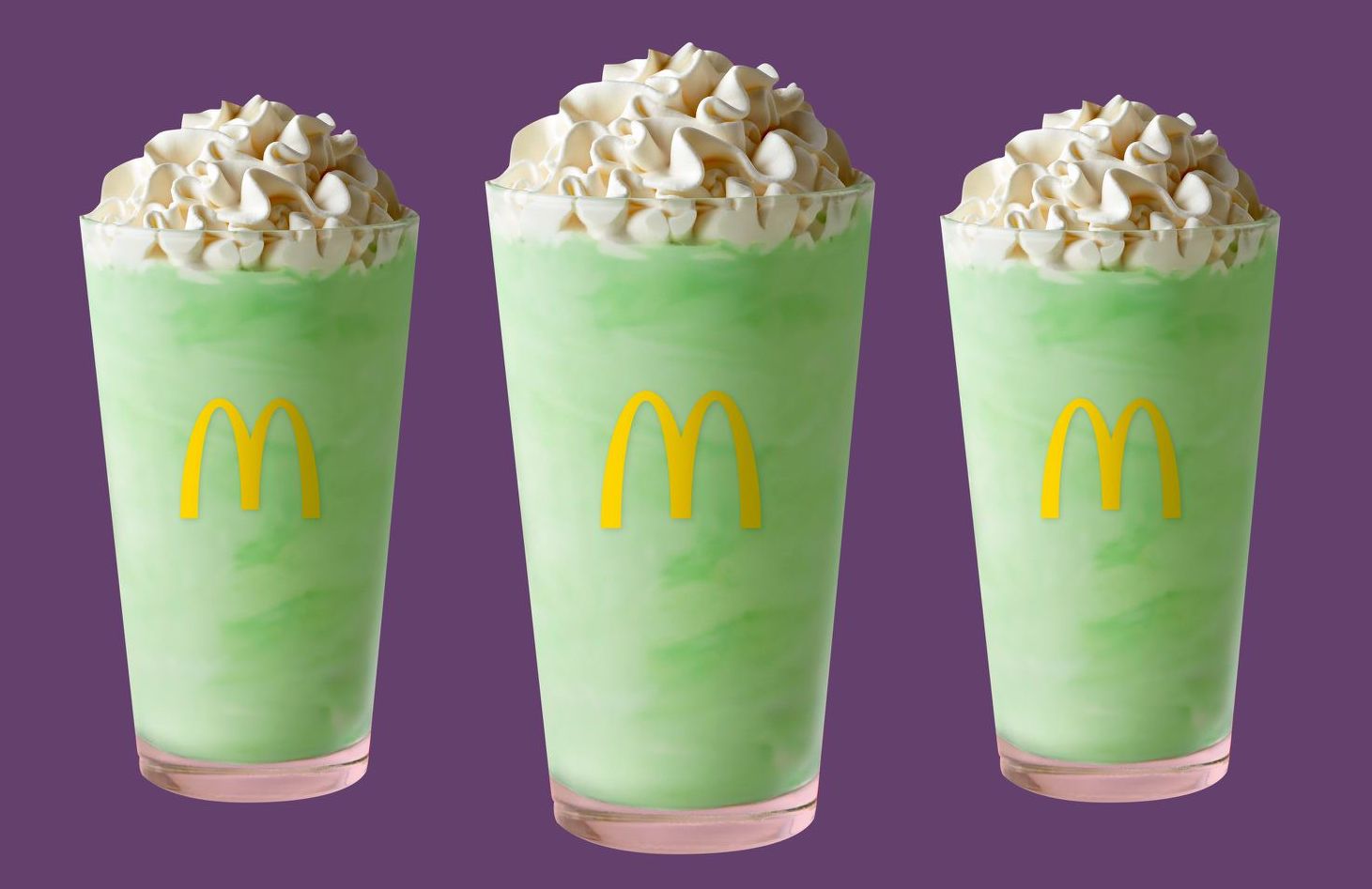 McDonalds-Shamrock-Shake-Is-Back-Already-FT-BLOG-0206-bbdc8c4ce5014e2294fd4a22a999943d - Social House News Three mint-green milkshakes topped with whipped cream, each with a McDonald’s logo on the cup against a purple background.