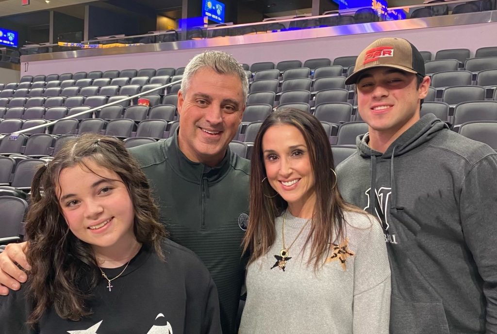 Four people pose together in a mostly empty stadium seating area, smiling at the camera.
