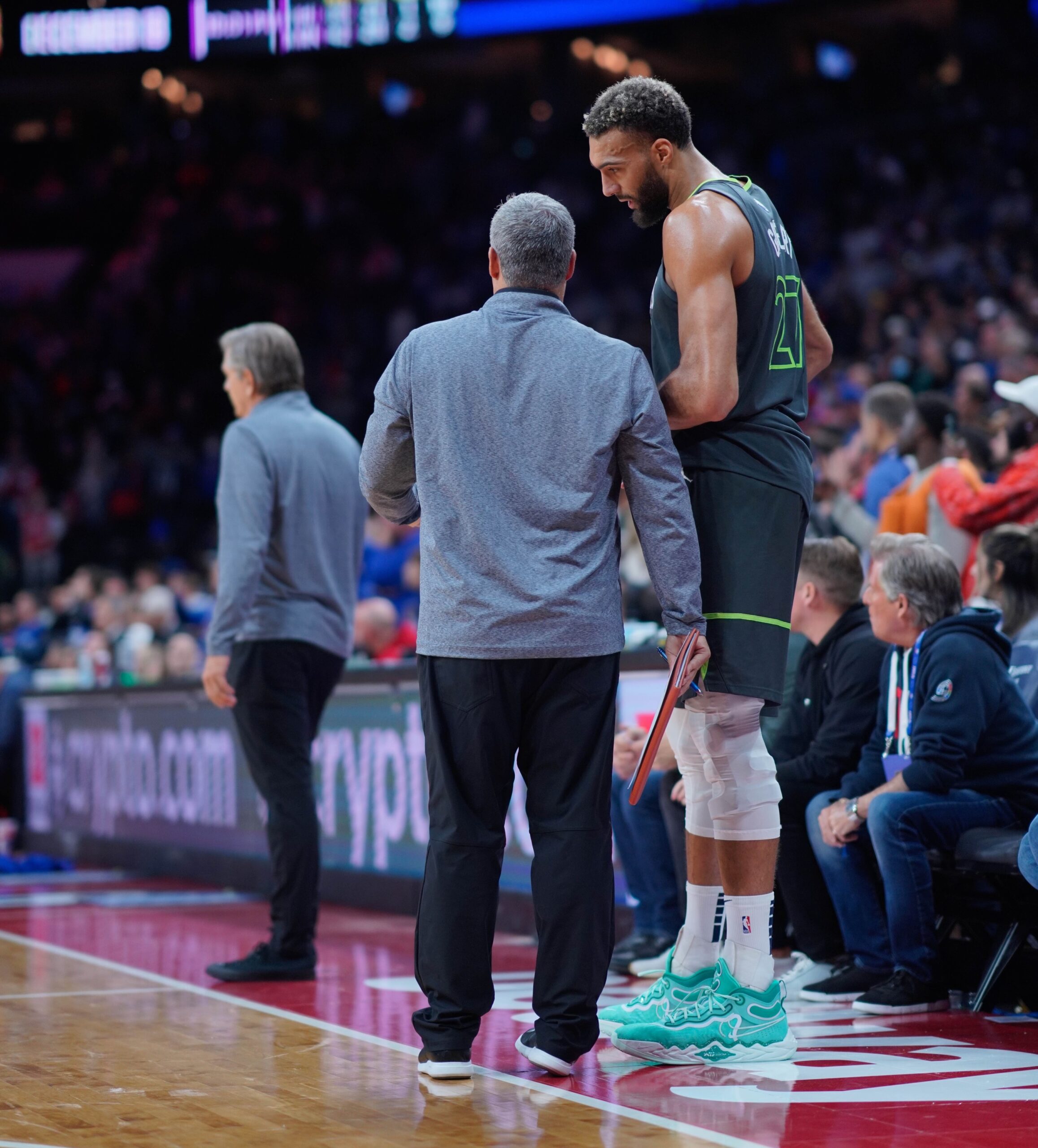 NBAjourney3-scaled - Social House News Basketball players and staff standing courtside during a game, one wearing teal sneakers and a leg wrap on his right leg.