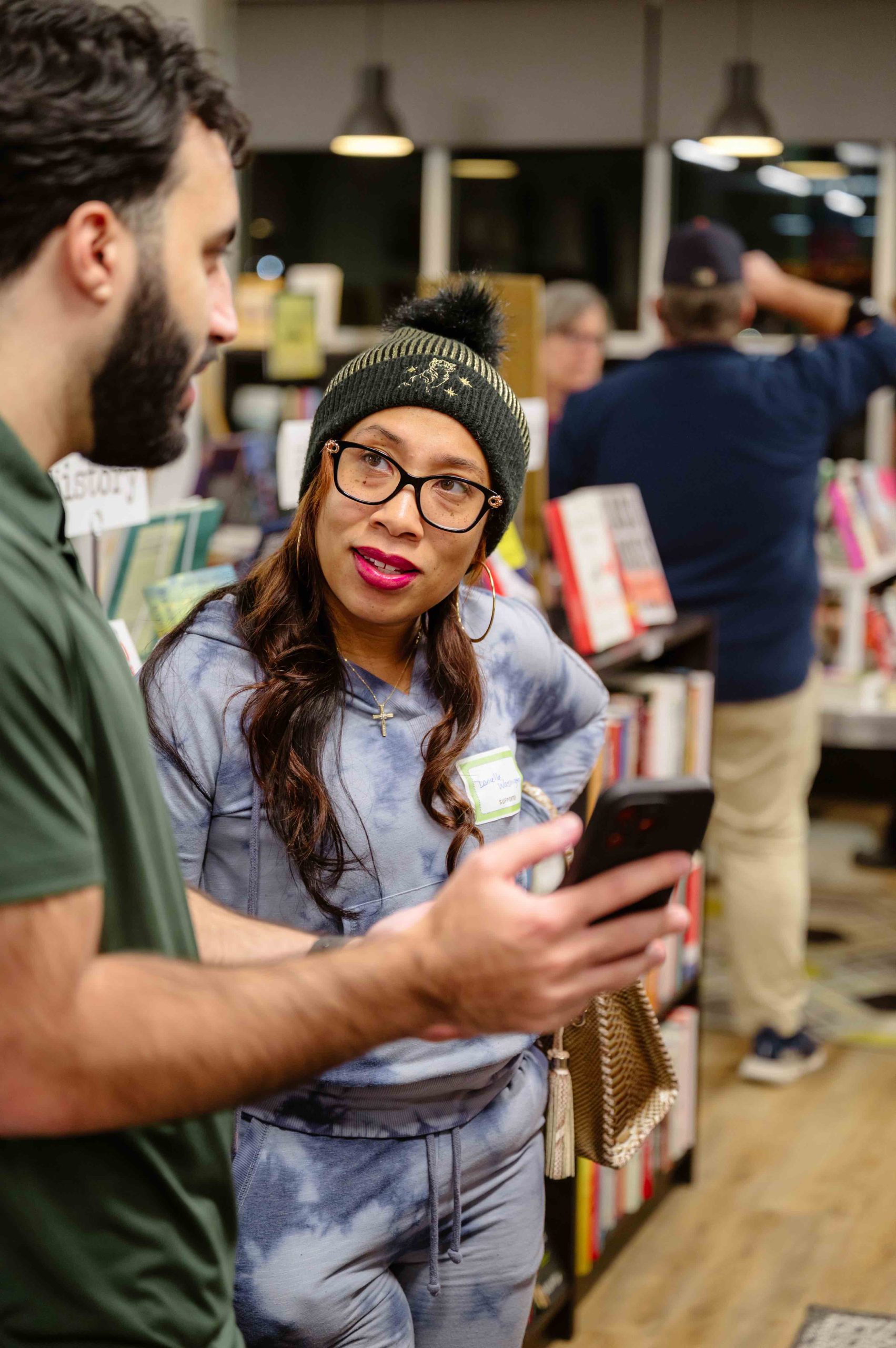Woman with glasses and a knit beanie talking to a man in a bookstore, holding a smartphone in her hand.