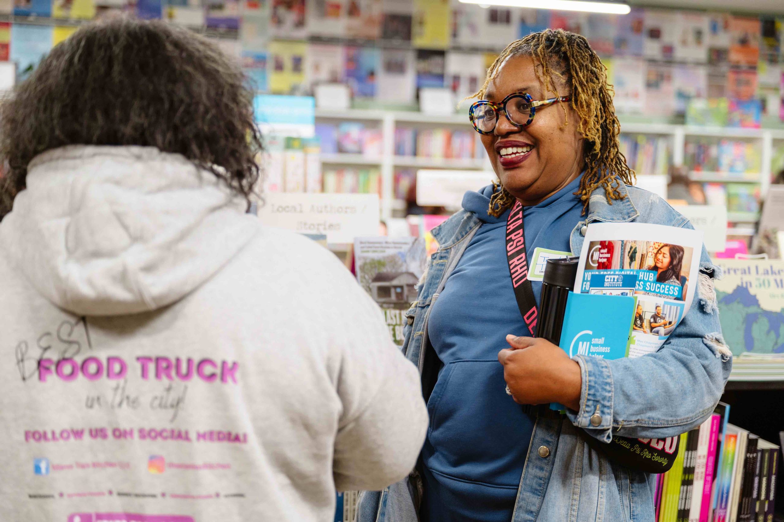A smiling woman with colorful glasses hands magazines to a customer in a bookstore aisle, shelves full of books behind them.