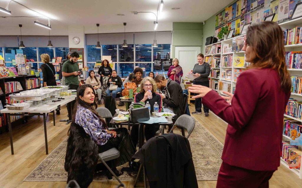 People sit at tables in a cozy bookstore, attending a community reading or meetup; shelves line the walls with colorful books nearby.