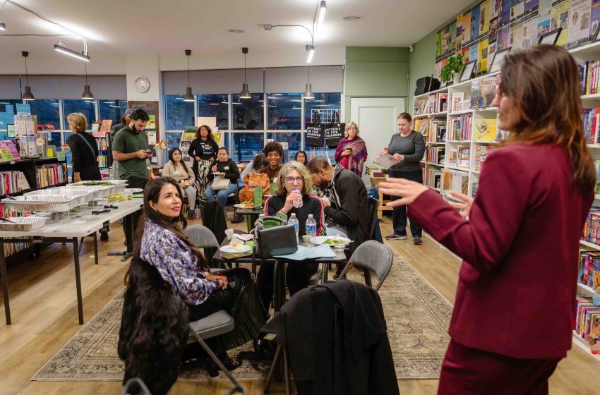 People sit at tables in a cozy bookstore, attending a community reading or meetup; shelves line the walls with colorful books nearby.
