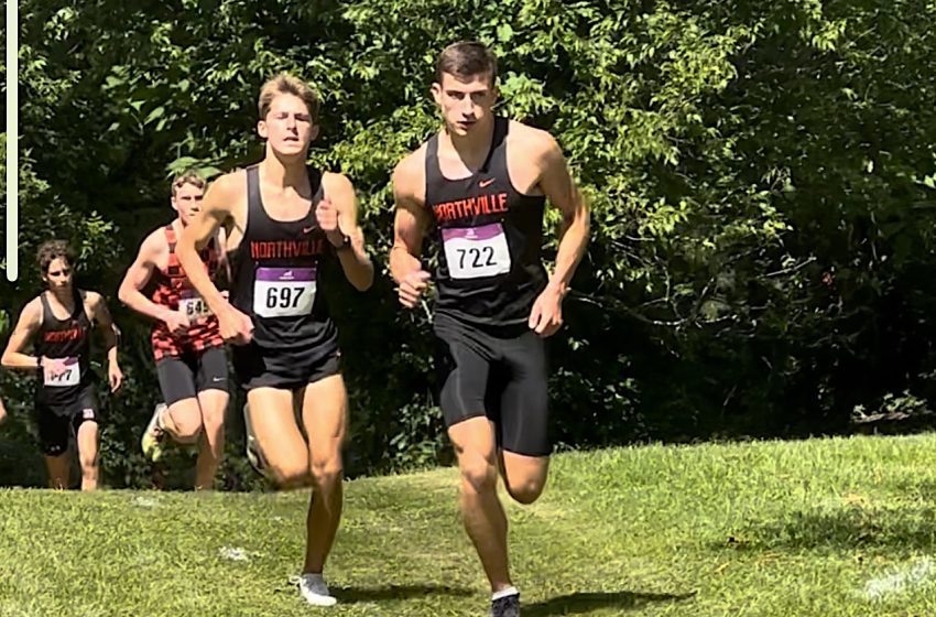 Young male cross-country runners in black Northville uniforms sprint uphill on a grassy course, bibs 697 and 722 visible with trees behind.