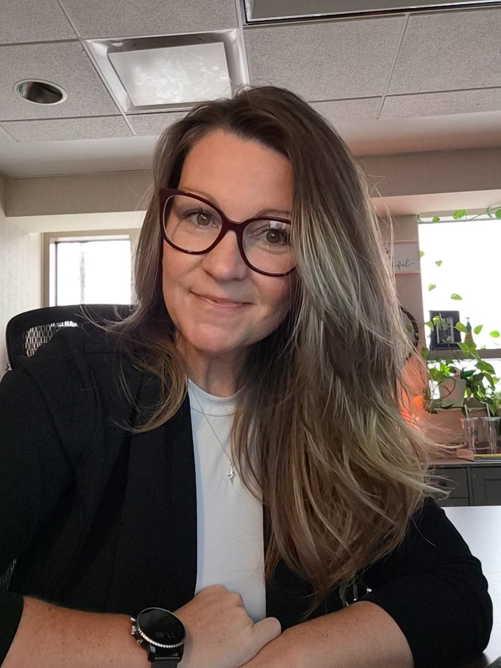 Photo Baker - Social House News Woman with long light brown hair and red glasses smiling at the camera in an office setting, wearing a white top and black blazer with a wristwatch nearby.