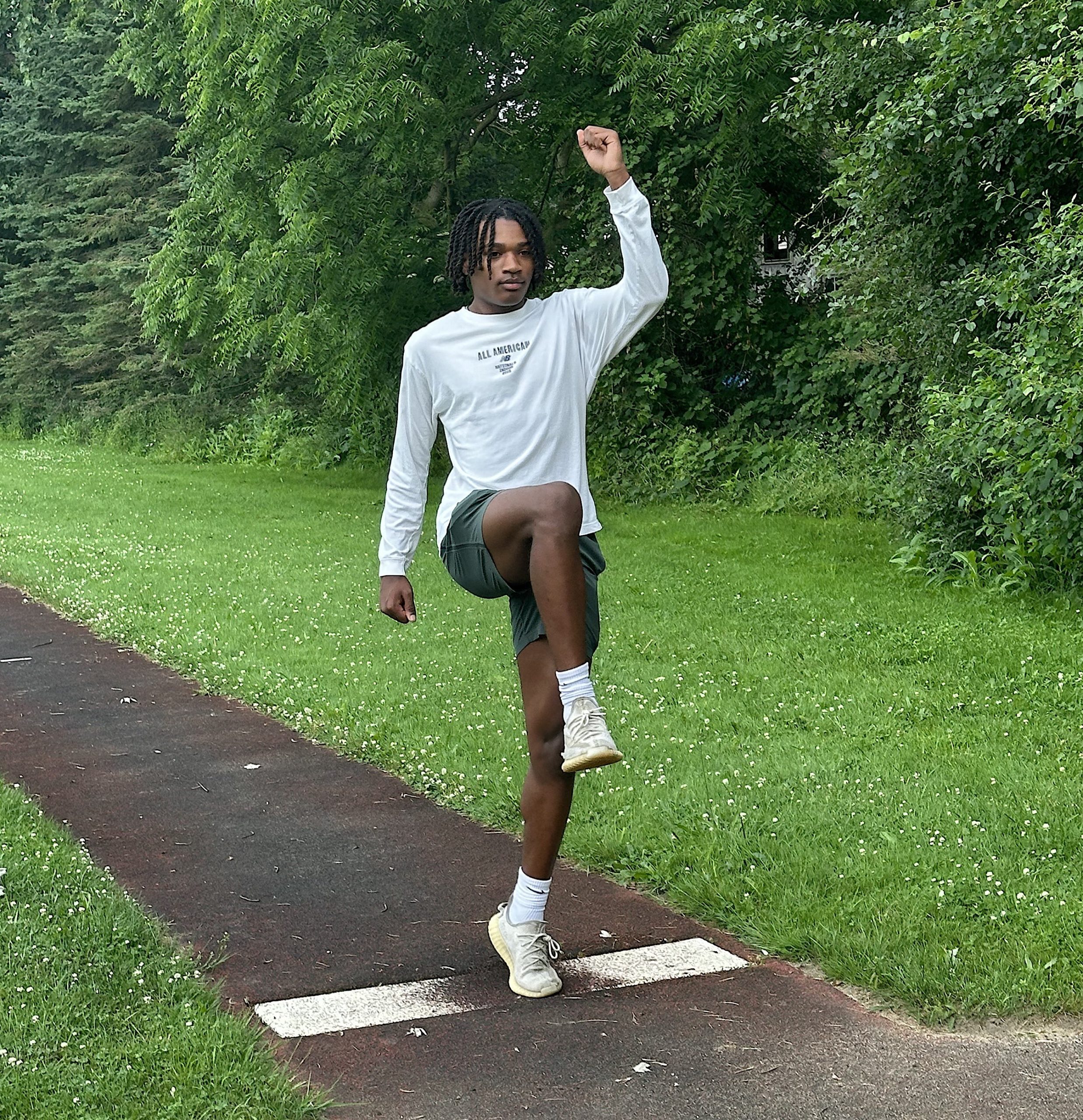 Young man in a white long-sleeve shirt leaps on a park path, left leg raised and fist raised, with green trees behind him.