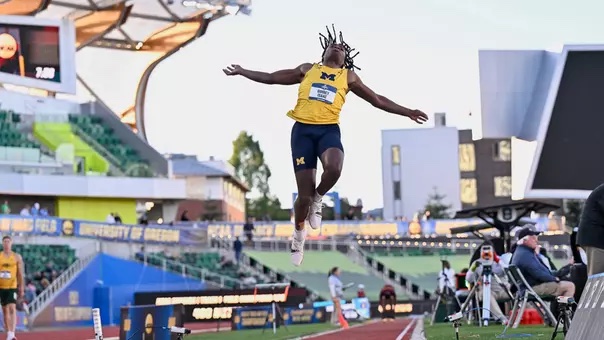 Male athlete in yellow Michigan uniform mid-air during a long jump at an outdoor track stadium.