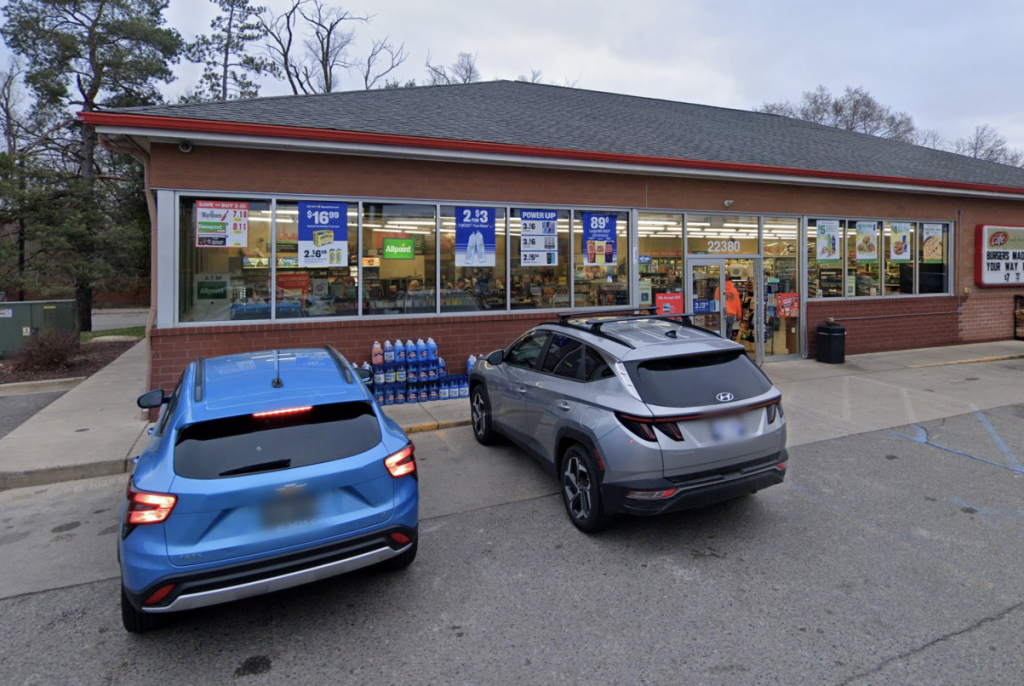 Two cars parked in front of a brick convenience store with large glass windows and promotional posters in the windows when approaching from the parking lot.