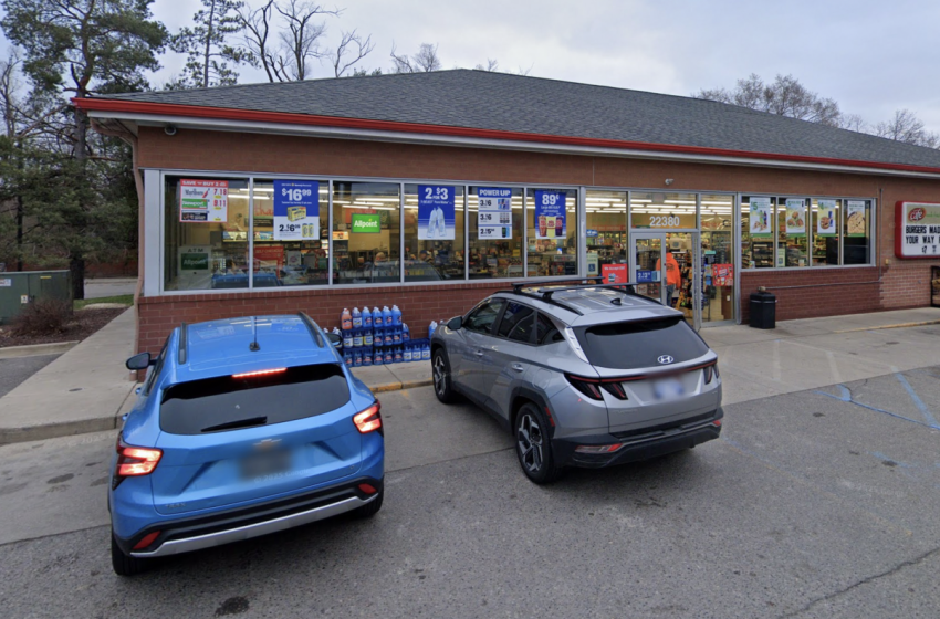 Two cars parked in front of a brick convenience store with large glass windows and promotional posters in the windows when approaching from the parking lot.
