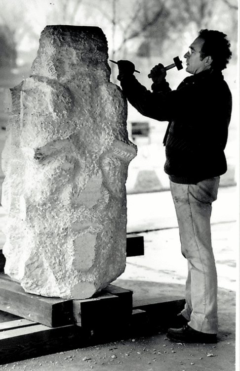 Screen Shot 2026-06-19 at 124637 PM - Social House News Man chisels a large rough stone sculpture outdoors with a hammer and chisel on a raised platform.
