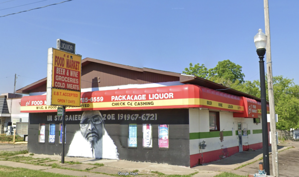 Liquor store on a street corner with a red-yellow awning, signs advertising beer, wine, and groceries, and a large mural of a man's face on the wall; blue sky above.