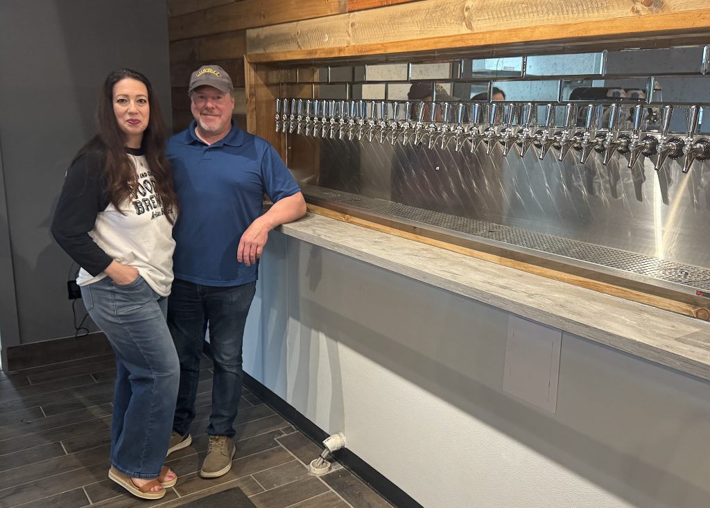 Two people stand beside a row of beer taps in a rustic taproom for a brewery photo.