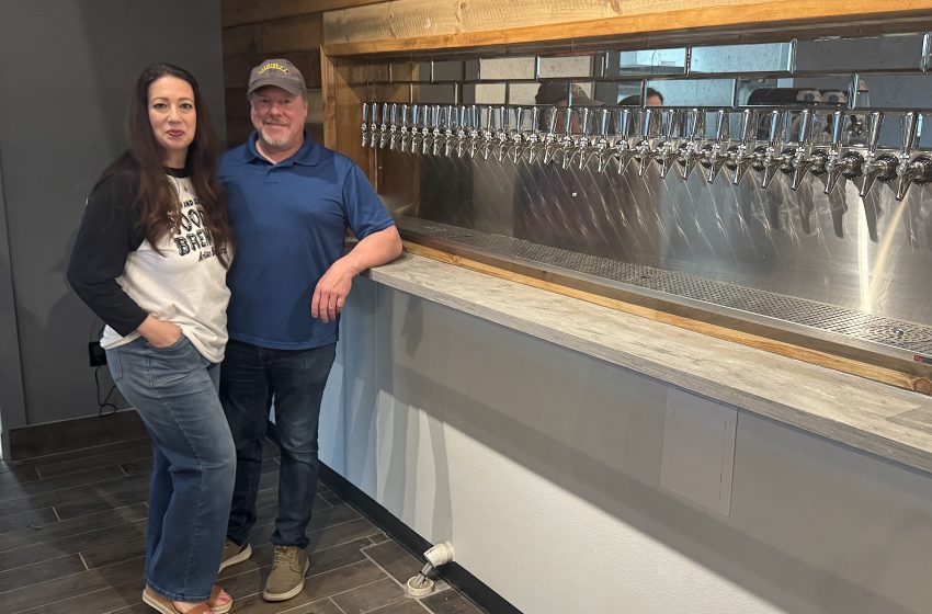 Two people stand beside a row of beer taps in a rustic taproom for a brewery photo.