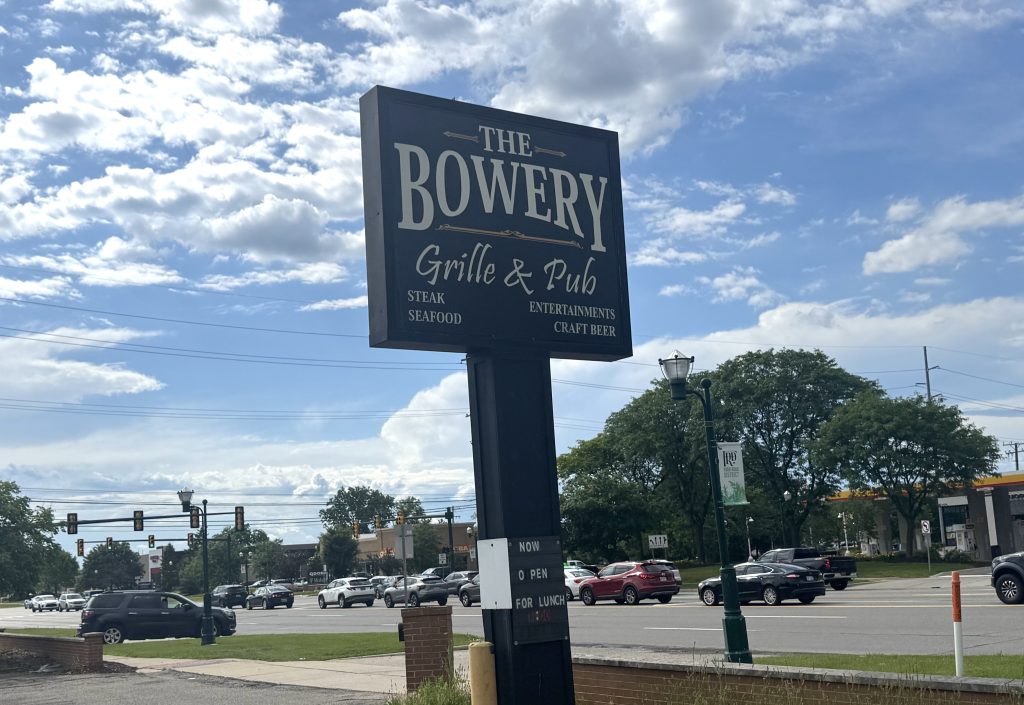 Tall blue sign for The Bowery Grille & Pub against a bright, partly cloudy sky with a parking lot and cars below.