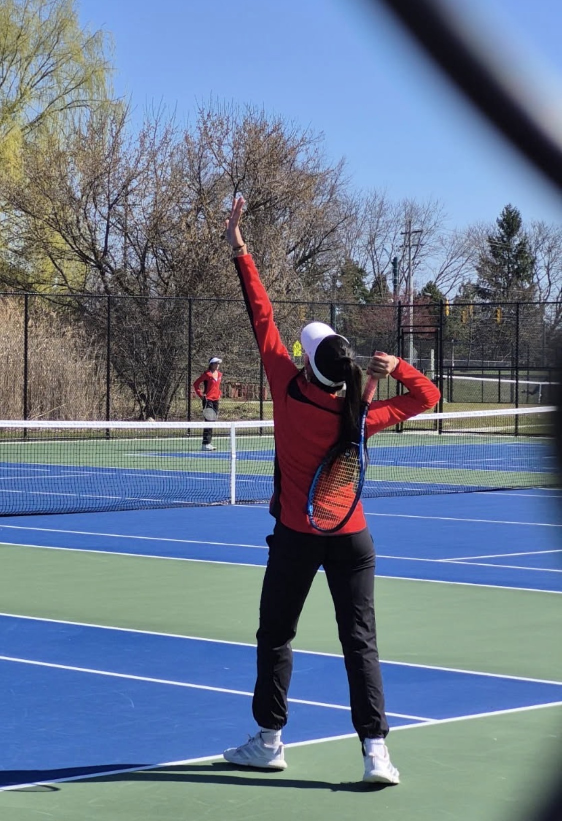 Screenshot - Social House News Tennis player in a red jacket serving on a blue and green court with a racket raised and one arm extended upward, outdoors on a sunny day.