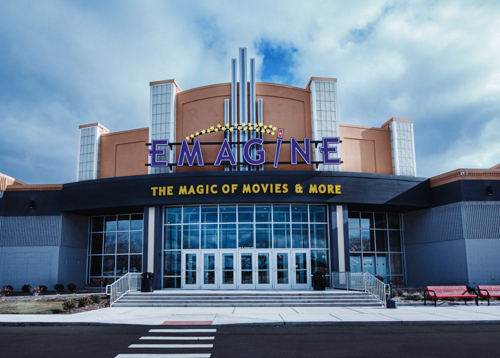 Front of Emagine cinema with a peach facade, purple sign, and glass doors against a blue, cloudy sky.