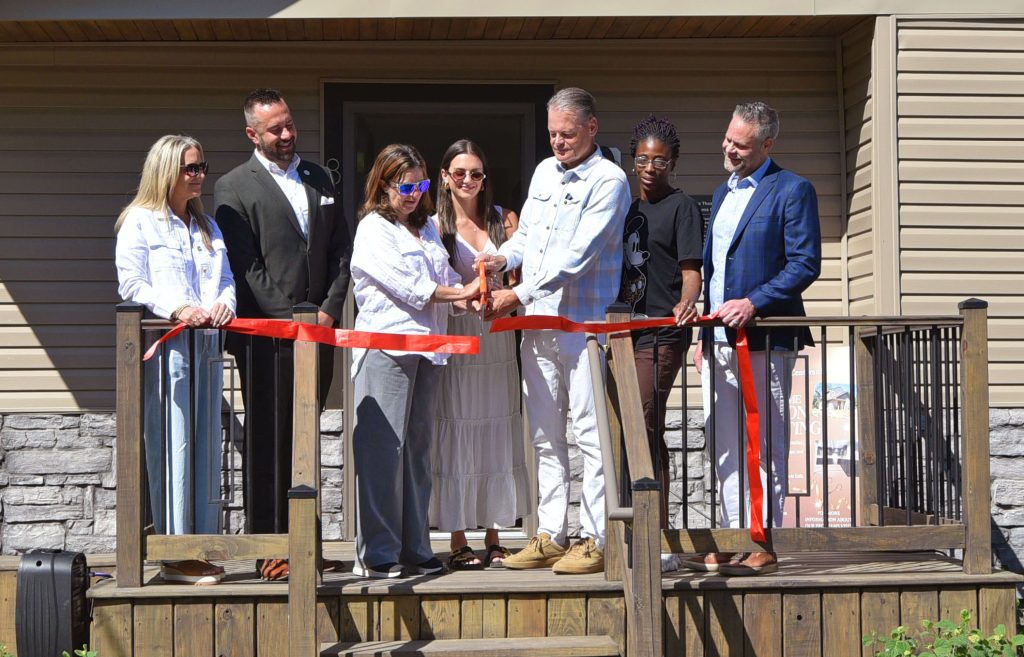 Seven adults stand on a wooden deck during a ribbon-cutting ceremony, hands on orange-handled scissors as they cut a red ribbon in front of a new building.