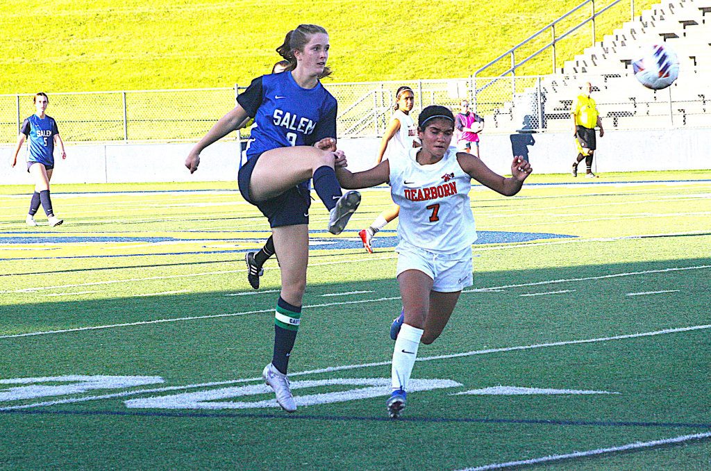 Girls' soccer action: a Salem player in blue jumps to kick the ball while a Dearborn player in white challenges nearby on a turf field.
