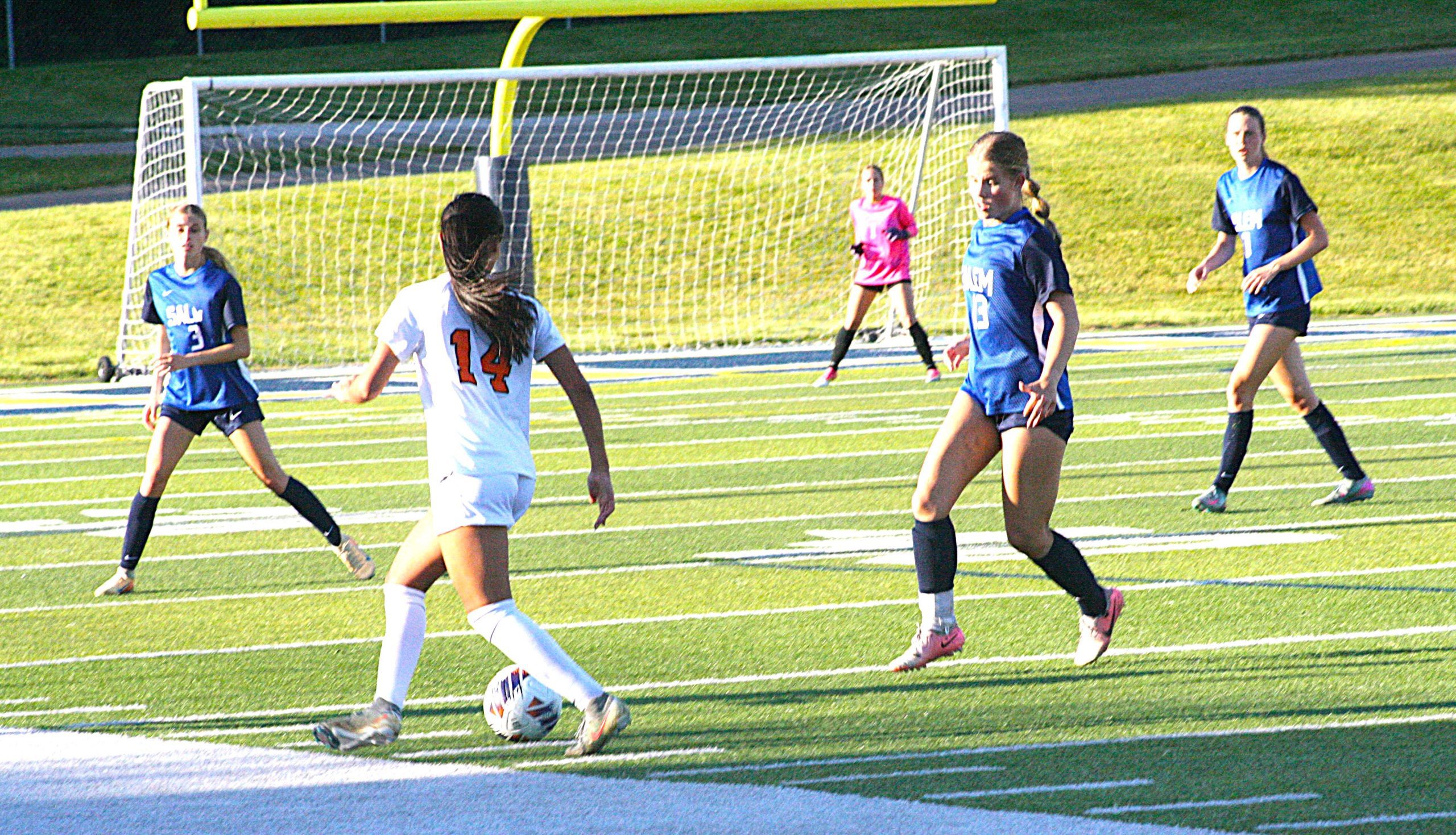 Girls' soccer game: a white-jersey player (#14) dribbles the ball toward the goal as blue-team players defend nearby.