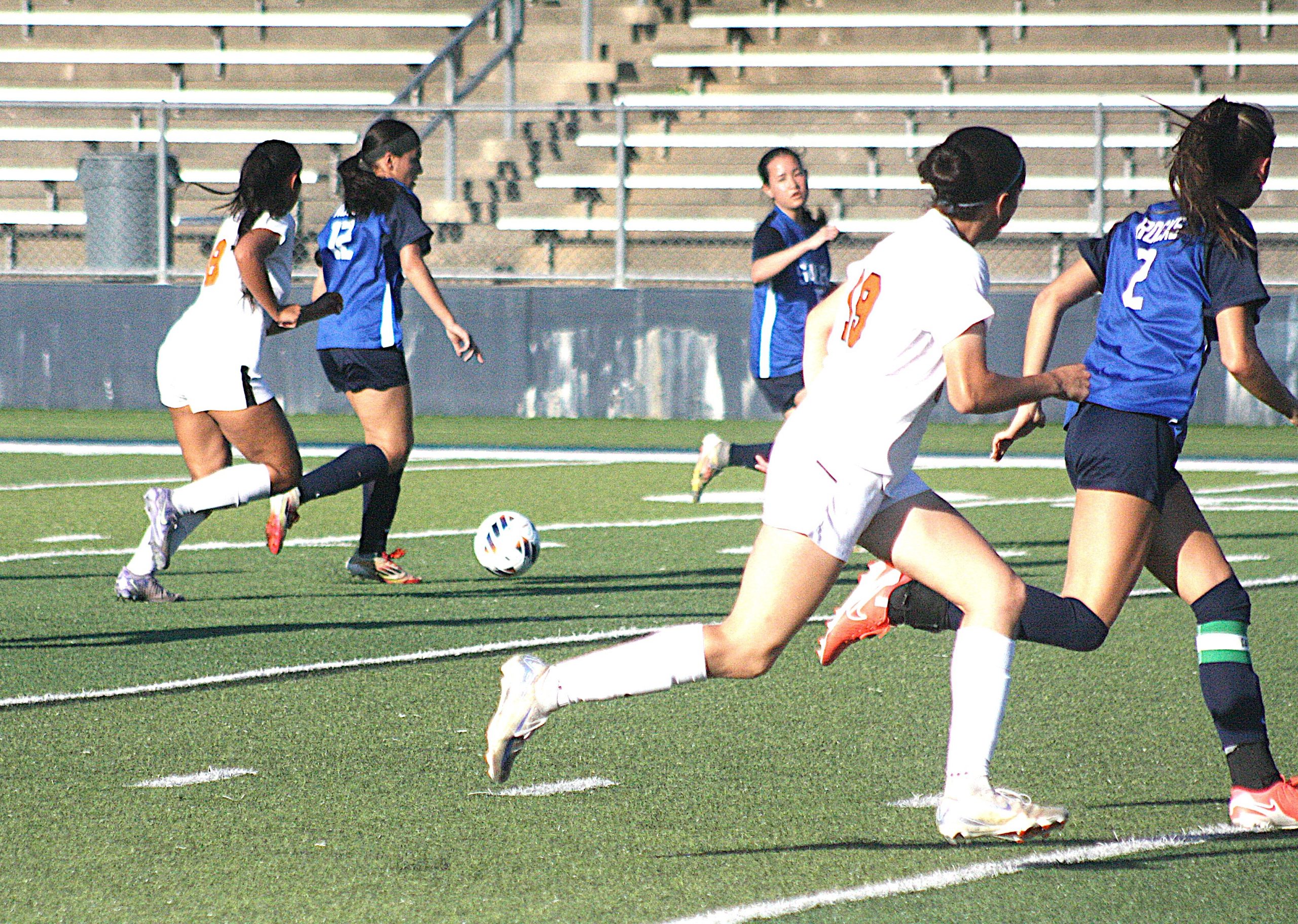 Girls' soccer match on a green turf field as players in blue and white uniforms chase the ball; bleachers in the background.