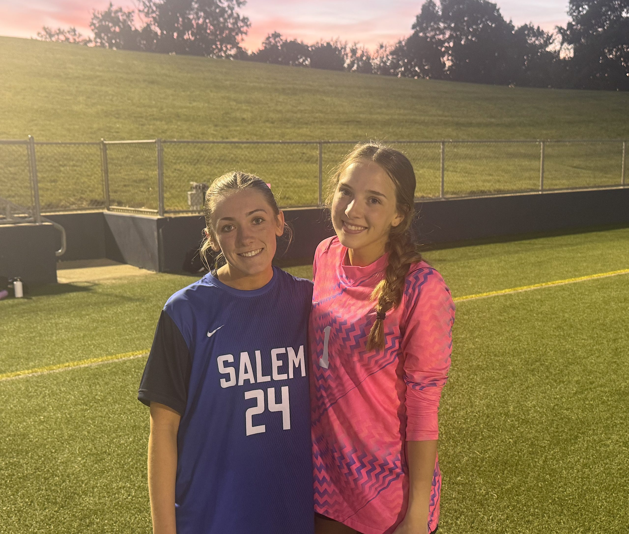 Two smiling female athletes pose together on a soccer field at dusk, one in a blue Salem jersey and the other in a pink patterned jersey.