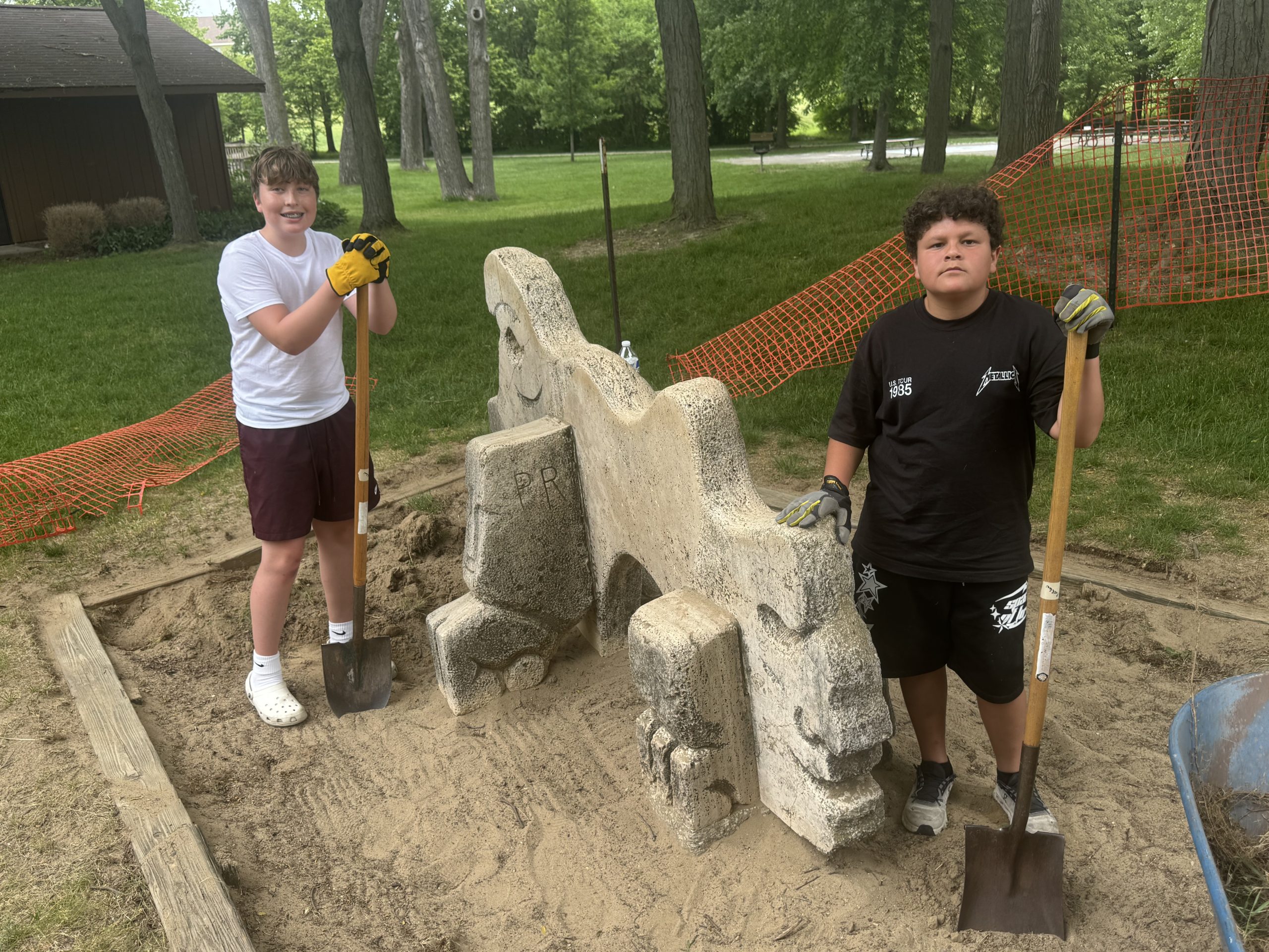 rockwell1 - Social House News Two boys wearing gloves and holding digging shovels stand beside a carved stone sculpture in a park, with orange construction mesh fencing behind them and trees in the background.