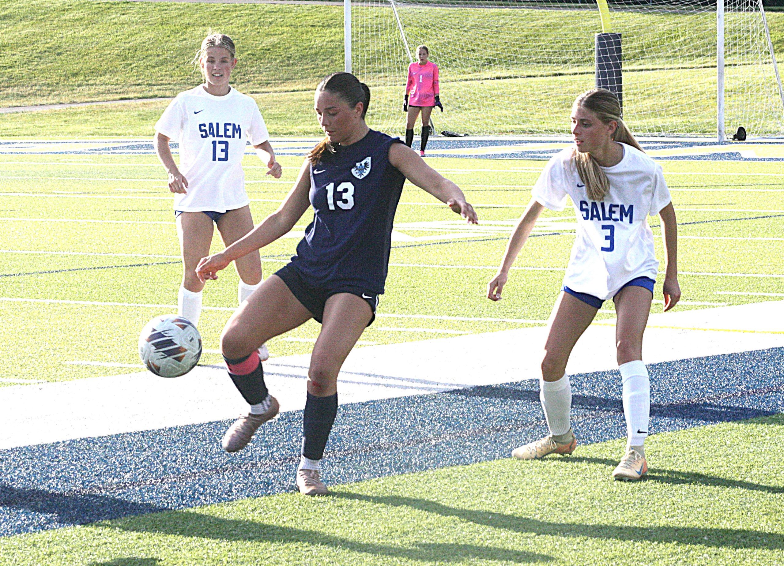 Girls' soccer match: player in navy jersey numbered 13 dribbles the ball while two Salem players in white jerseysClose by compete for possession on a sunny turf field with a goalkeeper in pink in the goal behind them.