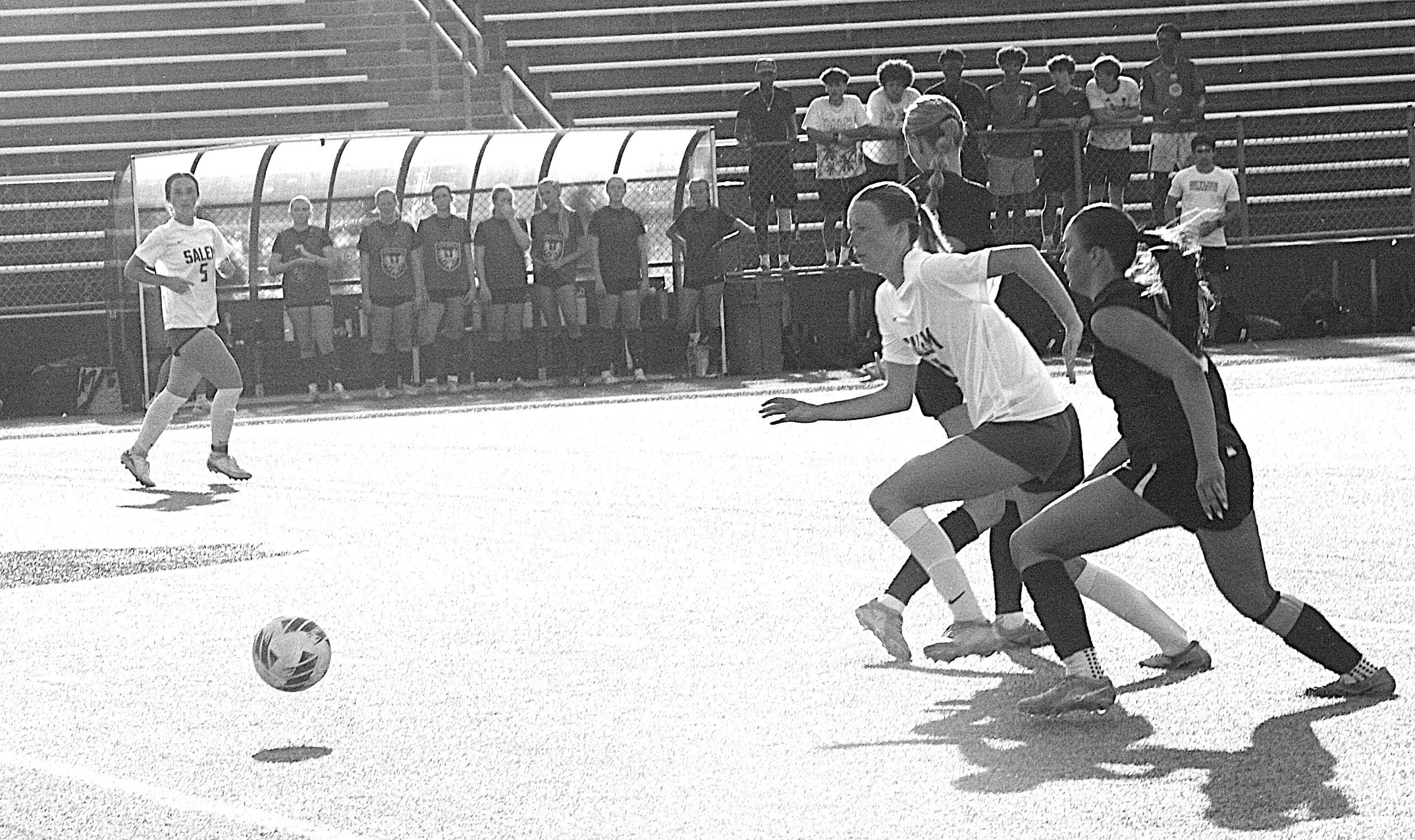 Soccer players sprint toward the ball on an outdoor field as spectators watch from the stands behind a bench area.
