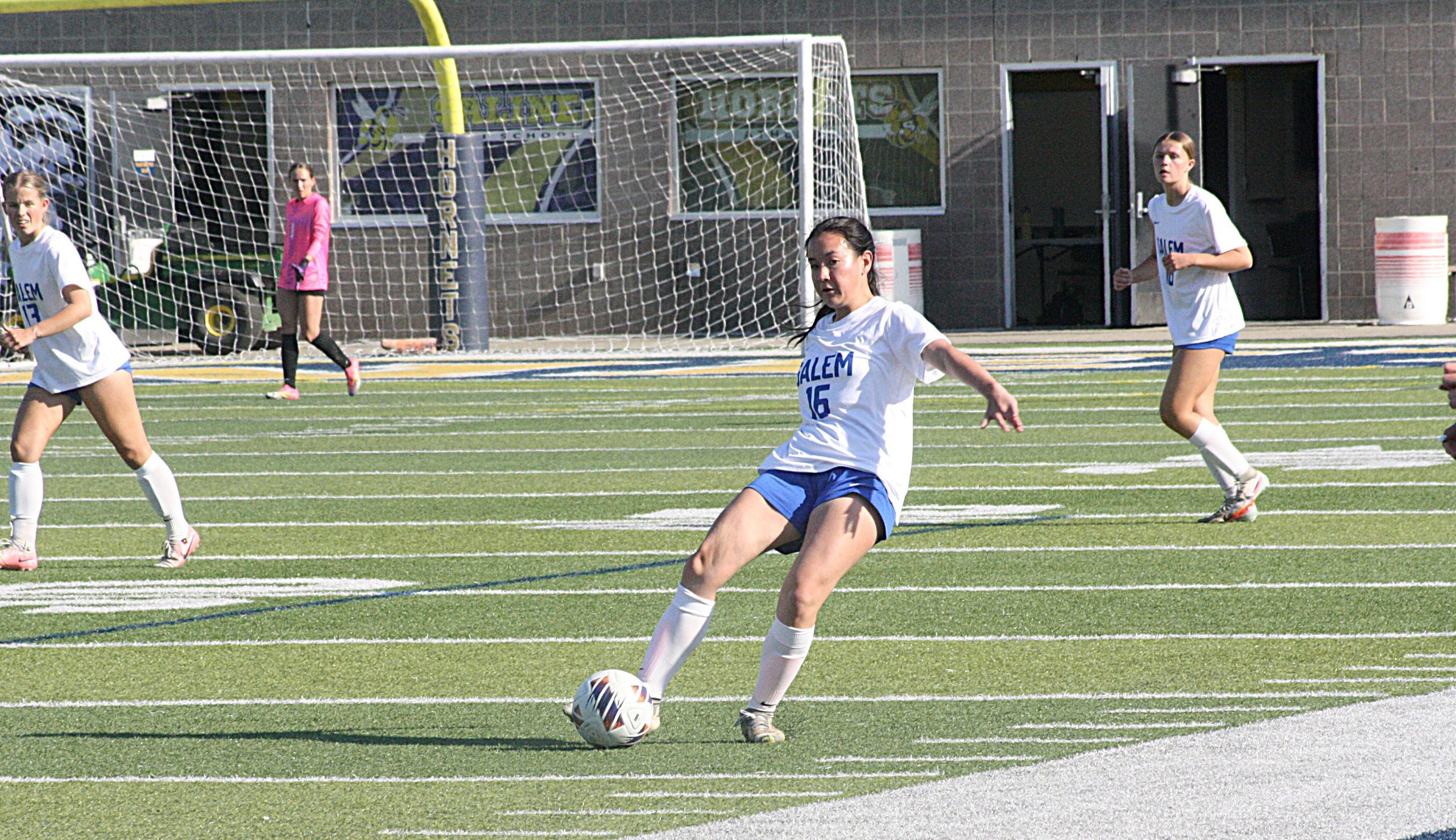 Soccer player in white Salem jersey number 15 kicking a ball during a game on a turf field with teammates and a goal in the background