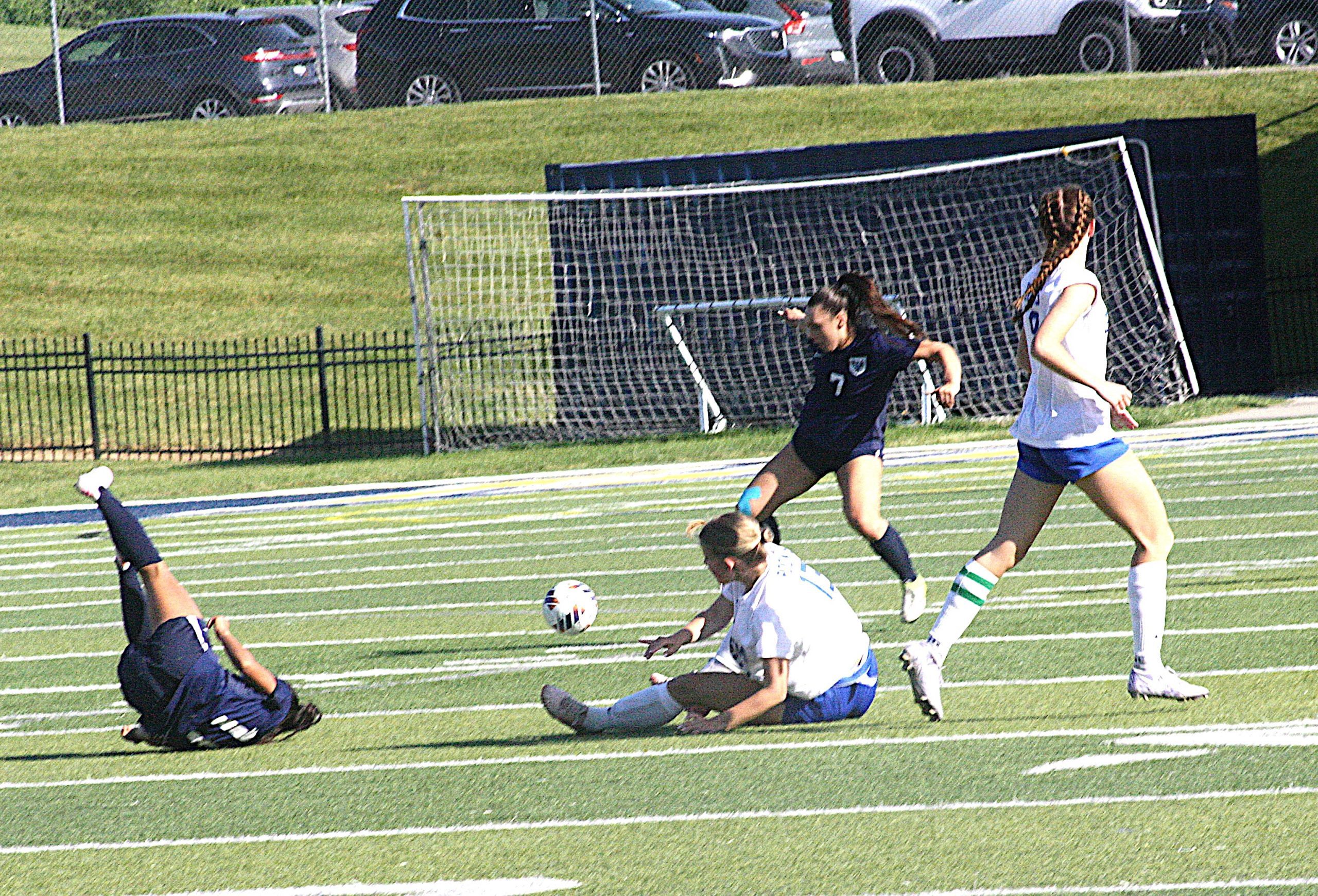 Soccer players chase the ball near the goal on a sunny field, one in navy and one in white jerseys amid a fallen opponent on the turf.