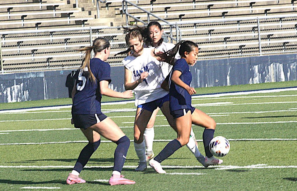 Female soccer players chase the ball during a match on a turf field with bleachers in the background.