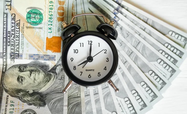 Analog alarm clock resting on a fan of US hundred-dollar bills, conveying the idea that time is money (time shown around 12:00).