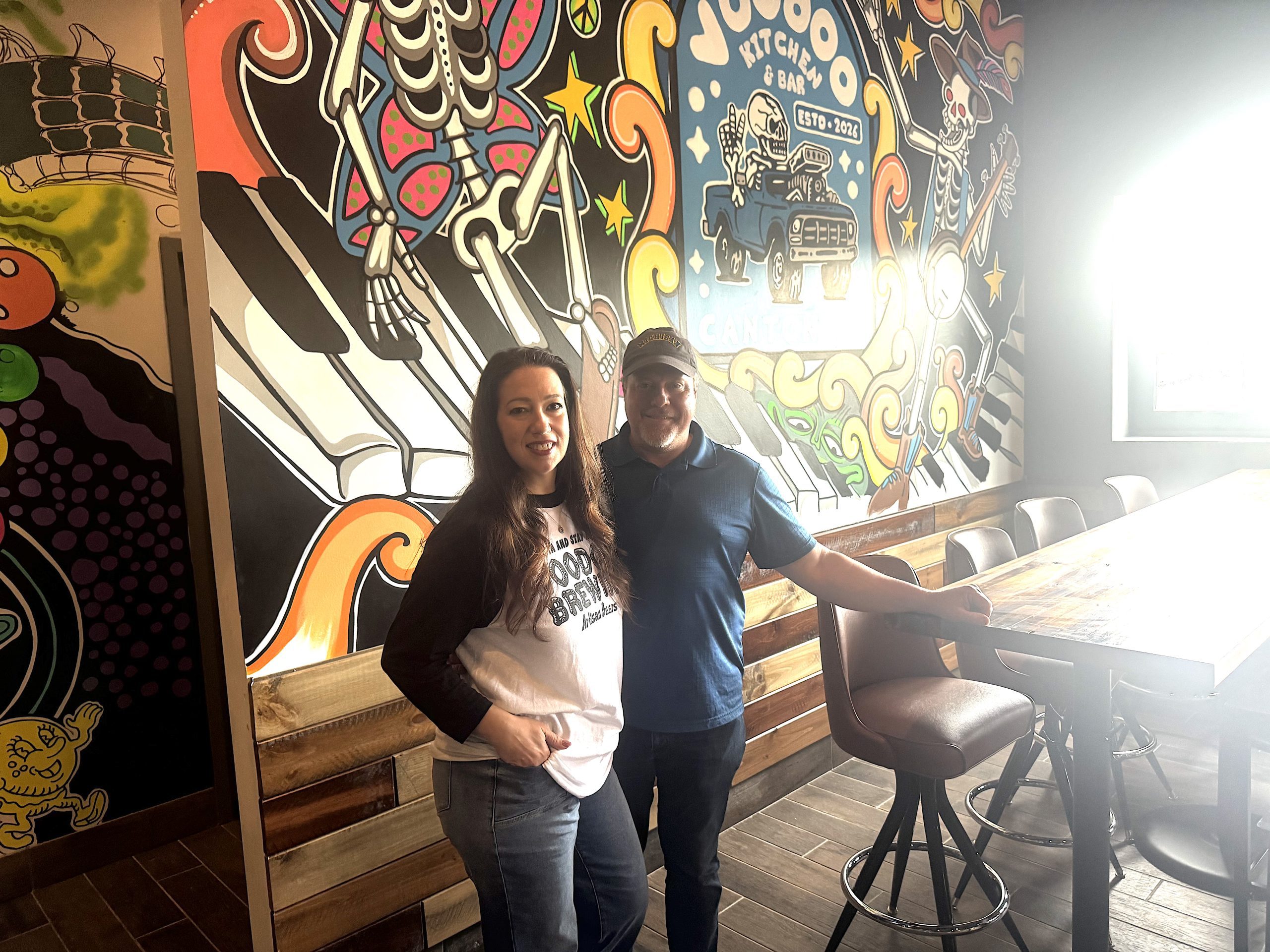 voodoo1 - Social House News Two people stand in front of a vibrant mural with guitars and whimsical skeleton art inside a cafe, smiling at the camera.